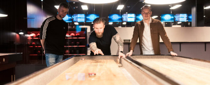 Krog med tre män som spelar shuffleboard inomhus, under moderna belysningsarmaturer och i en sportbar eller nöjeslokal, perfekt för sociala aktiviteter och tävlingar.