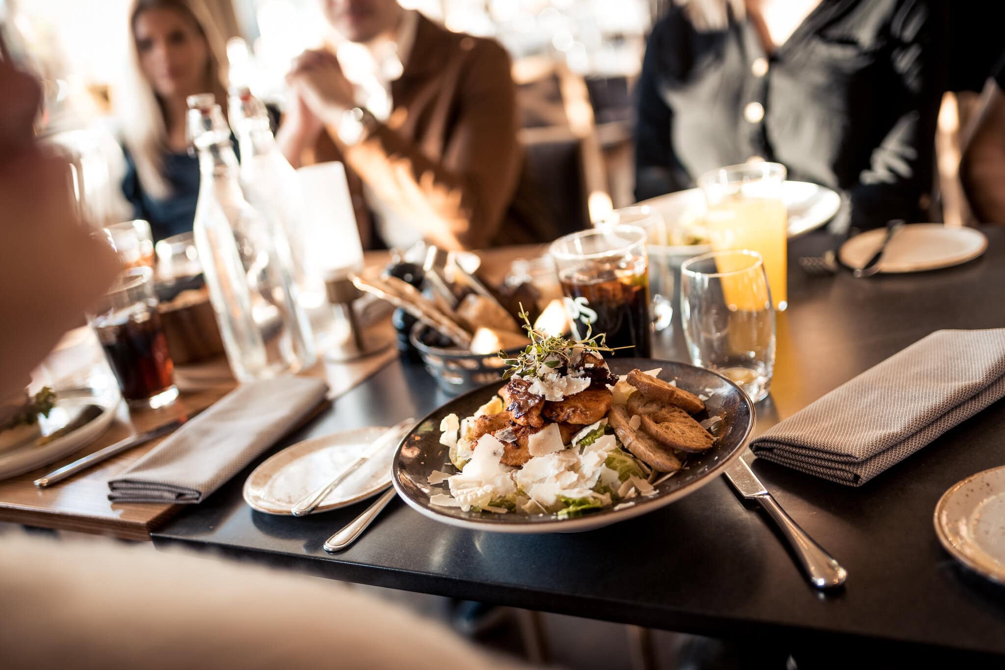 _DSC9395 Snyggt upplagt sallads- och antipastiplatta på restaurangbord med gäster i bakgrunden, fokus på mat och social kvällsmiddag.