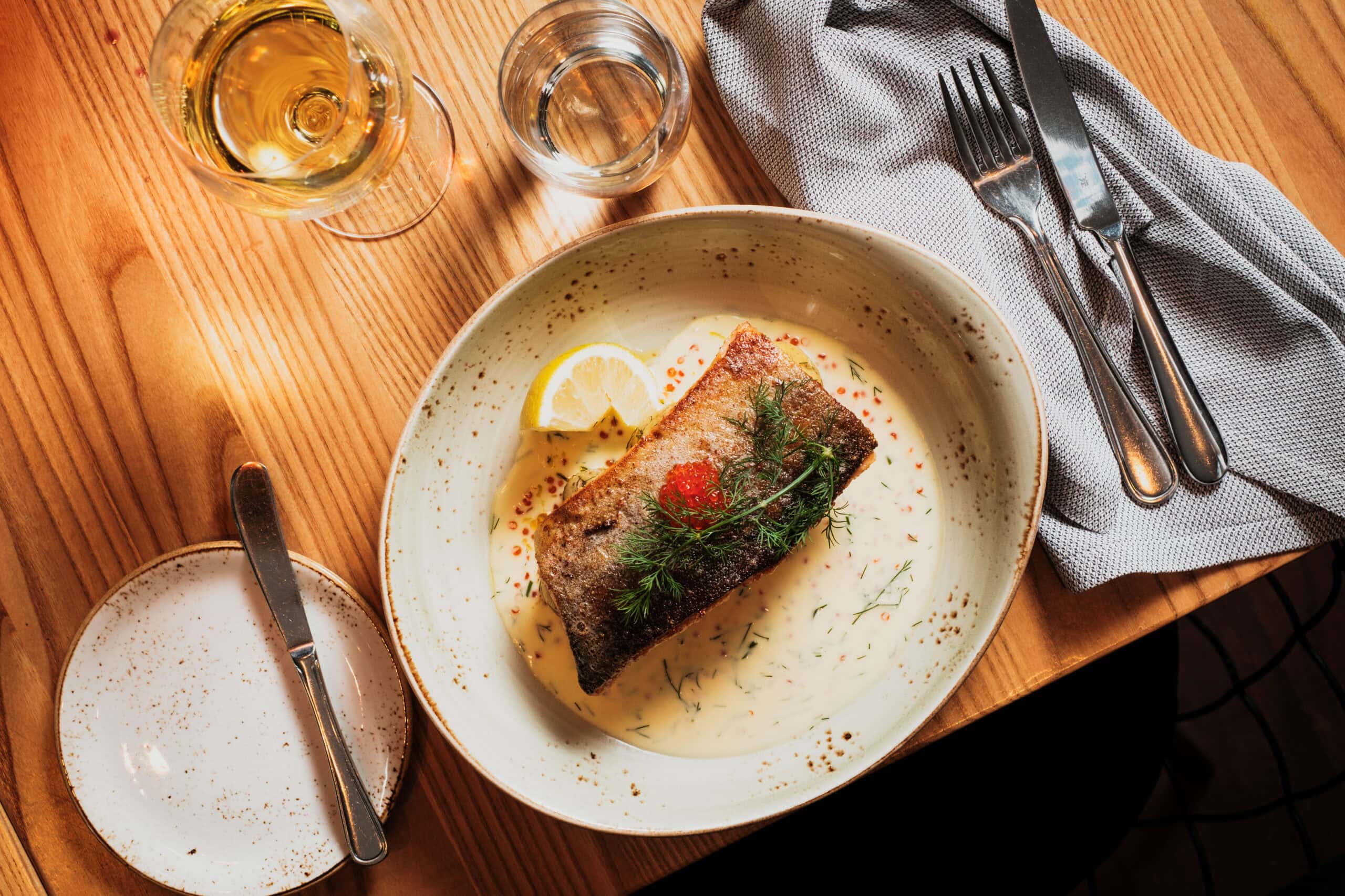 DSCF5544 Fiskrätt med citron, dill och saffranssås på ett träbord, serverad på en stor vit tallrik, med glas vin och vatten, perfekt för en mysig middag.