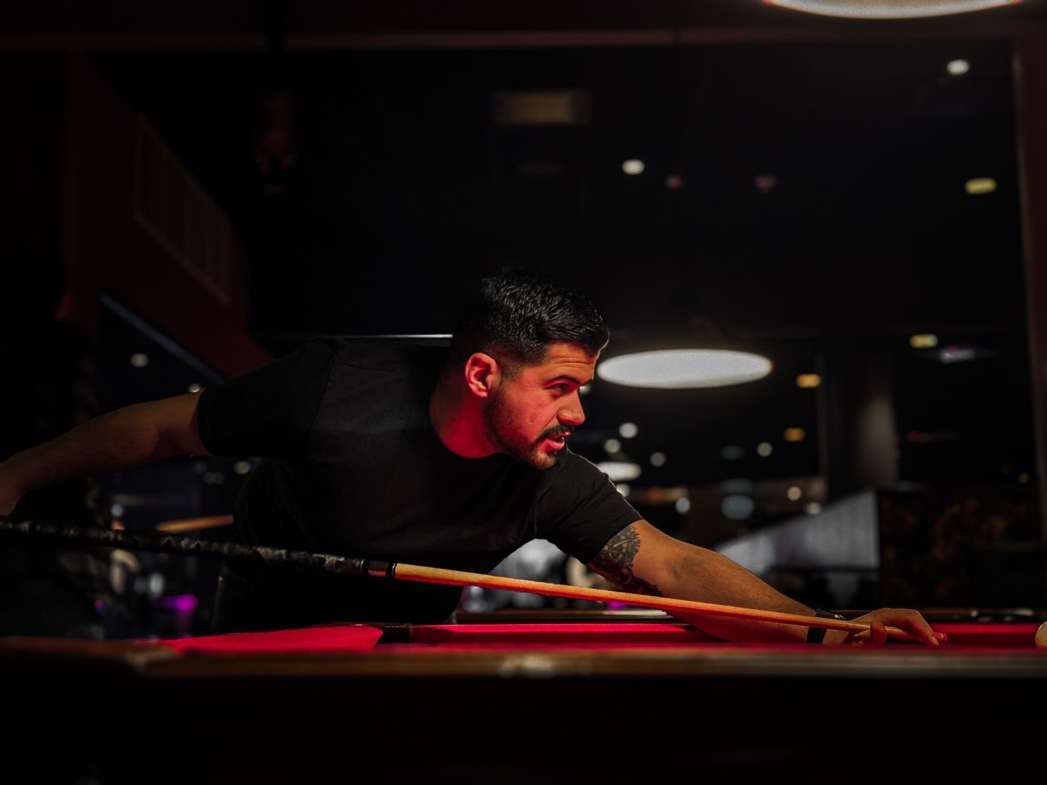 Onur biljard 2 Precis spelare som skjuter poolbollar vid ett biljardbord i en avslappnad bar eller spelhall Miljön är mörk med fokus på spelaren och belysningen ovanför poolbordet, perfekt för biljard och snookerälskare.